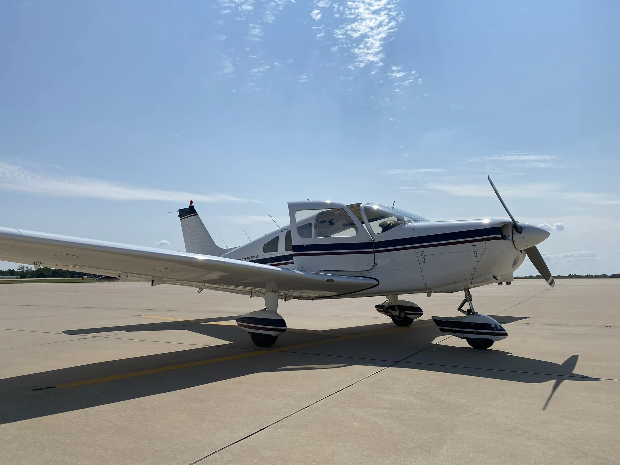 Piper Warrior N32204 on a sunlit airport ramp, viewed from the front quarter with its cabin door open